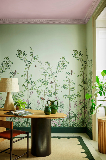 Green floral mural wallpaper featuring birds and butterflies (Bird & Bluebell - Pea Green) with a lamp and plant on a desk.