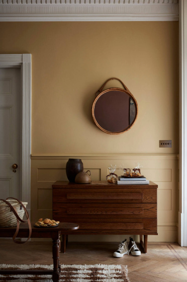 Hallway painted in muted gold shade 'Madeleine' with a wooden sidetable, round mirror and rug.