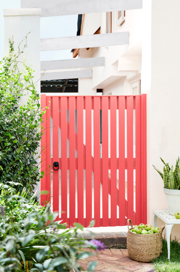 Coral red garden gate painted in 'Orange Aurora', alongside neutral white walls in 'Hollyhock', plants and bushes.