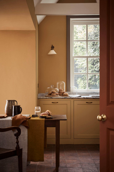 Kitchen space painted in honey shade 'Bombolone' with a paneled window, wooden table and chair.