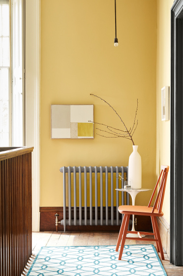 Hallway decorated with Light Gold walls, grey radiator, an orange chair, a patterned rug, and a white side table and vase.