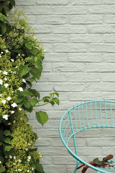 Exterior brick wall painted in grey green 'Normandy Grey' with a bush to the left and a turquoise blue chair to the right.