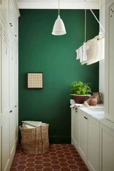 Laundry room painted in dark green shade 'Dark Brunswick' with a laundry basket, white cupboards and tiled floor. 