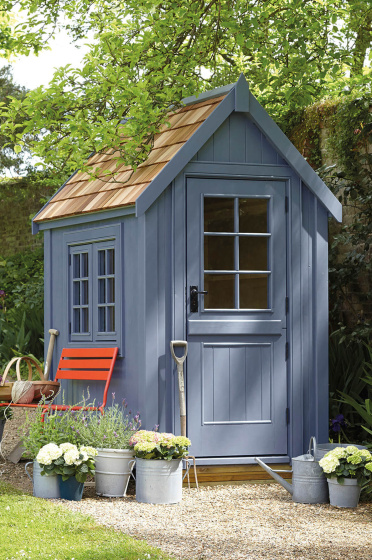 Outdoor shed painted in warm blue 'Juniper Ash' with a bright red chair surrounded by potted plants and trees.