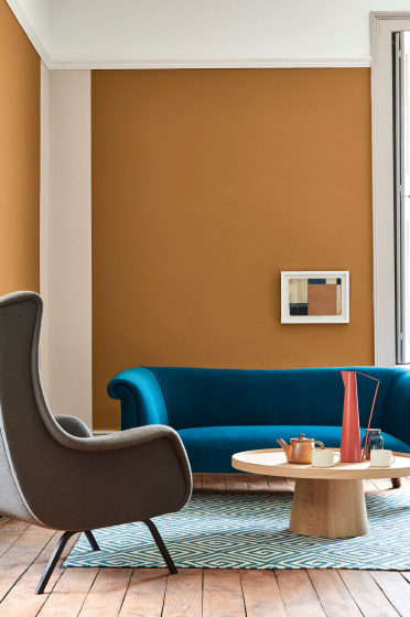 Living space painted in dark orange (Middle Buff) with a contrasting deep blue sofa, grey armchair and wooden table on a rug.
